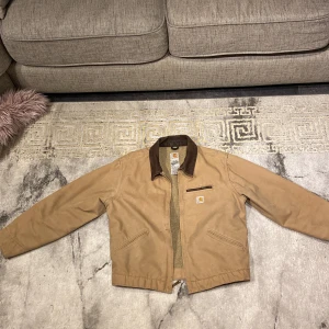 Vintage Carhartt Detroit jacka (model wj097) - Säljer en beige jacka från Carhartt med brunt krage och dragkedja. Jackan har en praktisk bröstficka med dragkedja och två sidofickor. Perfekt för höst och vår. Väldigt sällsynt jacka 