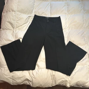 Svarta kostymbyxor från Pull&Bear - Snygga svarta kostymbyxor från Pull&Bear med en klassisk design. Byxorna har en rak passform och är perfekta för en stilren look. De har en knapp och dragkedja framtill. De har inga fickor.