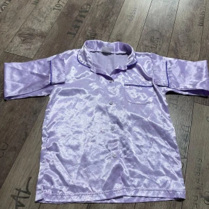 Lila satin pyjamas med hjärtmönster - Säljer en glansig lila pyjamas i satin med långa ärmar och knappar framtill. Blusen har ett subtilt hjärtmönster och en bröstficka med dekorativ kant. Perfekt för en stilfull och bekväm look. Obs större i storleken så passar utmärkt för någon som har xs- s. 