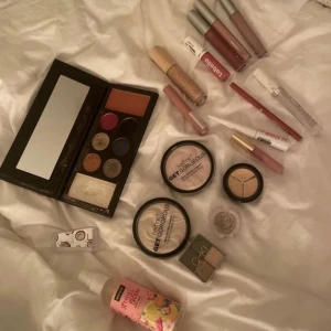 Samling av sminkprodukter - En samling sminkprodukter inklusive en ögonskuggspalett med flera färger, flera läppstift och läppglans i olika nyanser, en body mist, samt puder och concealer, highlight, handsprit och även eyeliner