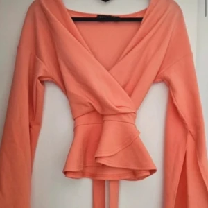 Persikofärgad omlottopp från Boohoo - Trendig persikofärgad omlottopp från Boohoo med långa ärmar och volangdetaljer. Toppen har ett knytband i midjan för en smickrande passform. Perfekt för att ge en färgklick till din garderob!