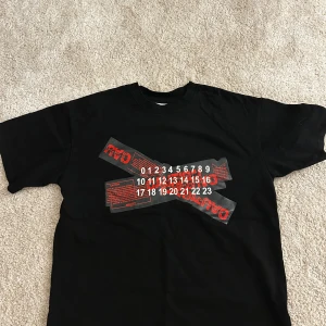 Svart t-shirt från Maison Margiela - Snygg svart t-shirt från Maison Margiela med unikt tryck i rött och vitt på framsidan. Perfekt för en stilren och modern look.