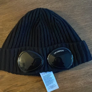 Svart ribbad mössa från C.P. Company - Snygg svart ribbad mössa från C.P. Company med unika glasögondetaljer framtill. Perfekt för att hålla stilen under kalla dagar.