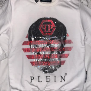 Vit tröja från Philipp Plein med paljettdetaljer - Säljer en vit tröja från Philipp Plein med en stor paljettutsmyckning på framsidan som föreställer en dödskalle och logotyp. Baksidan har tryck med text och dödskallemotiv. Tröjan har långa ärmar och en rund halsringning. Storlek L i junior så passar xs-s