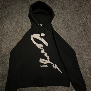 Svart hoodie från Kenzo - Säljer en svart hoodie från Kenzo med en stor vit logotyp och texten 'Paris' på framsidan. Hoodien har en klassisk design med dragsko i huvan och ribbade muddar vid ärmslut och nederkant. Riktigt fet hoodie som passar till det mesta! Tveka inte att höra av er för funderingar eller mer bilder! Priset är ej hugget i sten🙌🏻