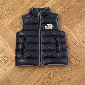 Mörkblå dunväst från Moncler - Snygg mörkblå dunväst från Moncler med dragkedja och två fickor framtill. Västen har en hög krage och ett märke på bröstet. Perfekt för kyligare dagar. Vi har strykt över ett namn inuti jackan! Den är i storlek 134!