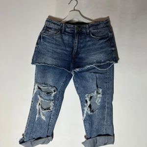 Blå jeans med slitningar från Zara - Säljer ett par coola blå jeans från Zara med slitna detaljer och upprullade benslut. Jeansen har en unik design med dubbla lager och slitningar på framsidan. Perfekt för en trendig look!🥰🥰🥰