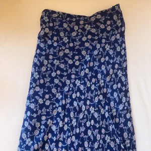 Blommig långkjol - Säljer en vacker blå långkjol med ett blommigt mönster i vitt och rosa. Kjolen har en volangdetalj vid fållen som ger en fin rörelse. Nytt skick🌸