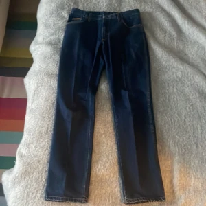 Mörkblå jeans från Wrangler 32x34 - Snygga mörkblå jeans från Wrangler med klassisk femficksdesign och märkets logga på bakfickan. Jeansen har en rak passform och är perfekta för en avslappnad stil. De har en knappgylf och bälteshällor för extra komfort. W32 L34. Riktigt fint skick!