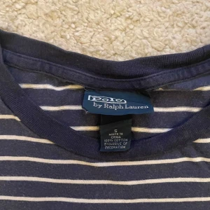 Randig t-shirt från Polo Ralph Lauren - Säljer en snygg randig t-shirt från Polo Ralph Lauren i marinblått och vitt. T-shirten är kortärmad och har det klassiska Polo-logotypen broderad i gult på bröstet. Perfekt för en avslappnad stil.