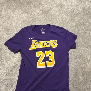 Lila Lakers t-shirt från Nike - Säljer en lila Lakers t-shirt från Nike med nummer 23 och texten 'Lakers' i gult på framsidan. Baksidan har namnet 'James' och nummer 23 i gult. Perfekt för basketfans!
