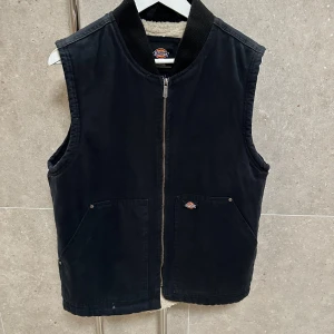 Svart väst från Dickies - Säljer en svart väst från Dickies med dragkedja framtill. Västen har en enkel design med två fickor och en liten logotyp på bröstet. Perfekt för lager-på-lager stil. Skönt foder på insidan som håller dig varm för en kall vår dag!