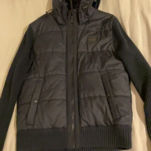 Svart jacka/cardigan från BOSS - Säljer en snygg svart jacka/cardigan från BOSS med quiltad framsida och stickade ärmar. Jackan har en dragkedja framtill och en praktisk huva. Den är tunn så perfekt för våren och sena sommar kvällar. Inga difunktioner. Pris kan såklart förhandlas. Köpte från Zalando. Vid frågor så kontakta bara👍🏼