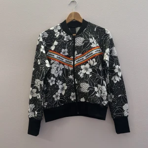 Blommig tjock tröja från Adidas - Snygg bomberjacka från Adidas med ett blommigt mönster i svart och vitt. Jackan har en dragkedja framtill och ribbade muddar vid ärmslut och nederkant. Perfekt för att ge din outfit en cool touch! är i storlek l men är väldigt liten hade kunnat passa en s eller m 