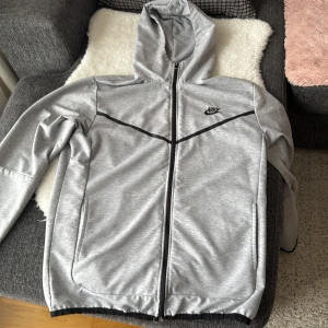 Grå huvtröja från Nike tech - Säljer en grå huvtröja från Nike tech med dragkedja och svart logga på bröstet. Tröjan har långa ärmar och en praktisk huva. Perfekt för en sportig look eller avslappnade dagar. Priset kan diskuteras vid snabb affär 