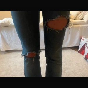 Blå jeans med slitningar - Säljer ett par blå jeans med slitningar på knäna. De har en bootcut-stil och är lågmidjade. Perfekta för en avslappnad look.