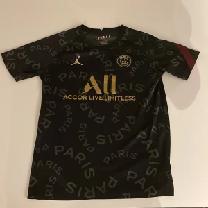Svart Limited Edition PSG fotbollströja från Jordan - Snygg svart Limited Edition PSG fotbollströja med guldiga detaljer från Jordan. Tröjan har ett mönster med texten 'PARIS' över hela. PSG-loggan och Jordan-loggan syns tydligt på framsidan. Perfekt för alla fotbollsfans!