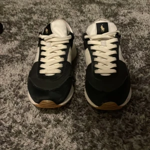 Ralph Lauren Trainers - Snygga svarta och vita sneakers från Polo med klassisk design. Skorna har en vit sula med brunt mönster undertill och är dekorerade med Polo-loggan på sidan och tungan. Perfekta för en stilren look. Sitter väldigt skönt på foten och skulle säga att de passar för storlek 38-39.