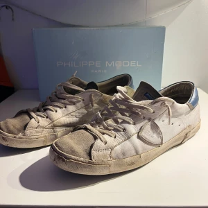 Vita sneakers från Philippe Model - Säljer ett par vita sneakers från Philippe Model med blå detaljer på hälen. Skorna har en klassisk design med snörning. Perfekta för en avslappnad stil. Storlek 44
