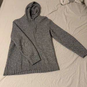 Grå stickad hoodie - Mysig grå stickad hoodie med långa ärmar och ribbade muddar. Perfekt för kyliga dagar med sin bekväma passform och klassiska design. Huvan har snören för justering.grish! Materialet är inte som ny men funkar bra. Priset kan diskuteras