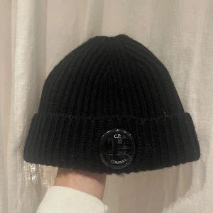 Svart ribbad mössa från C.P. Company - Snygg svart ribbad mössa från C.P. Company med en rund logodetalj framtill. Perfekt för att hålla värmen under kyliga dagar. Mössan är i en klassisk design som passar till det mesta. Använd max två gånger, dock har y:et försvunnit lite men inget man lägger märke till!