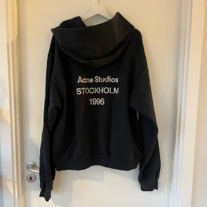 Svart hoodie från Acne Studios - Säljer en svart hoodie från Acne Studios med texten 'Stockholm 1996' på ryggen. Tröjan är tillverkad i en bomullsblandning och har en klassisk passform med långa ärmar och en bekväm huva. Perfekt för en stilren och avslappnad look.