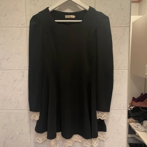 Svart klänning med spetsdetaljer - Elegant svart klänning från The Pretty Dress Company med långa ärmar och vackra vita spetsdetaljer vid ärmslut och nederkant. Klänningen har en snörning i ryggen som ger en fin passform.