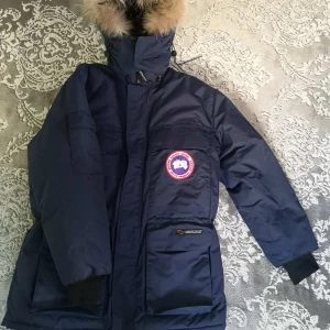 Blå dunjacka från Canada Goose - Säljer en blå dunjacka från Canada Goose med pälsdetaljer på huvan. Jackan har en broderad logotyp på bröstet. Den är i storlek M, men är stor i storlek så passar L. Används bara några gånger.