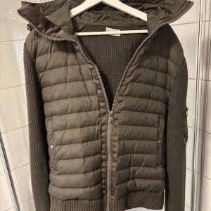 Olivgrön cardigan från Moncler - Säljer en olivgrön quiltad cardigan från Moncler med stickade ärmar och en unik färg. Jackan har en dragkedja framtill och en praktisk ficka med dragkedja på ärmen. Köptes från Plick för ett år sen runt 3200kr och säljes nu billigt.