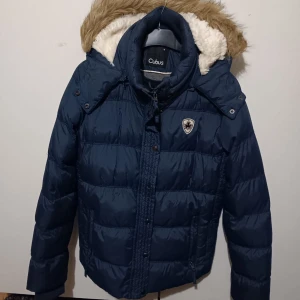 Mörkblå dunjacka från Cubus - Säljer en snygg mörkblå dunjacka från Cubus med avtagbar huva och fuskpäls. Jackan har dragkedja och knappar framtill, samt ett emblem på bröstet. Perfekt för kalla vinterdagar! 🧥❄️