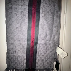 Grå Gucci  halsduk med ränder - Säljer en snygg grå Gucci halsduk med ett diskret mönster och breda ränder i rött och svart. Perfekt för att hålla sig varm och stilren under kyliga dagar. Den har fransar i ändarna som ger en extra touch. Passar både till vardags och festliga tillfällen.fick denna i present där av inga kvitton eller äktahets intyg där av garanterar jag inget men är väldigt säker på att den är äkta, så de mer upp till en själv skriv till mig för mer info