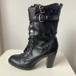 Svarta boots med klack och snörning - Snygga svarta boots med hög klack och snörning framtill. De har också dragkedja på sidan och två spännen upptill för extra stil. Perfekta för höst och vinter, och ger en cool look till vilken outfit som helst. Använd dem för en edgy stil på festen eller bara till vardags.