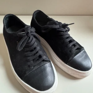 Svarta sneakers från Axel Arigato - Snygga svarta sneakers från Axel Arigato i mocka och skinn. De har en stilren design med snörning och en vit sula som ger en cool kontrast. Perfekta för både vardag och festliga tillfällen. Skorna är i bra skick och redo för nya äventyr! Strl 36
