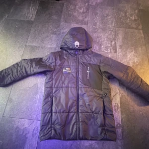 Svart jacka från Sail Racing size 170 - Säljer en svart jacka från Sail Racing i mycket bra skick. Den har en cool design med dragkedja och en praktisk huva. Perfekt för kalla dagar med sin quiltade stil. Jackan har också snygga detaljer som en bröstficka med logga och en inneretikett. Passar perfekt för höst och vinter!