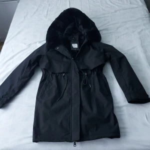 Svart parkajacka från CC Fashion Paris - Säljer en snygg svart parkajacka från CC Fashion Paris i storlek L. Jackan har en mysig fuskpälsfodrad huva och dragsko i midjan för en justerbar passform. Perfekt för kalla höst- och vinterdagar. Den är i nytt skick och har både dragkedja och knappar framtill.