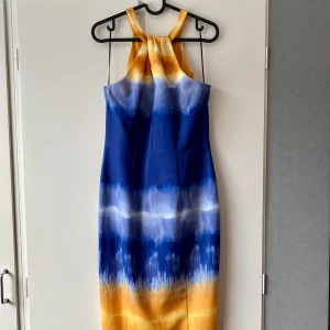 Blå och gul klänning med halterneck - Snygg halterneck-klänning i blått och gult med ett coolt mönster. Klänningen har en smickrande passform och är i mycket bra skick. Den är ärmlös och har en dragkedja i ryggen för enkel påklädning, samt en slits i bak. 