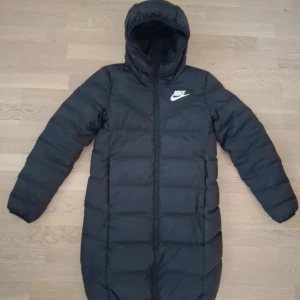 Svart och vit dunjacka från Nike - Säljer en snygg och varm dunjacka från Nike i svart och vitt. Jackan har en lång modell med huva och dragkedja framtill. Perfekt för kalla vinterdagar! Den är i mycket bra skick och redo för nya äventyr. Passar både till vardags och sportiga aktiviteter. Om du har andra frågor är det bara att skicka ett meddelande 