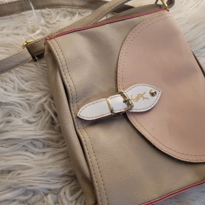 Beige och rosa axelväska med spänne - Säljer en snygg axelväska i beige och rosa med ett dekorativt spänne framtill. Väskan har en justerbar axelrem och en praktisk dragkedja upptill. Perfekt för vardagsbruk eller en dag på stan. Den är i mycket bra skick och redo för en ny ägare!