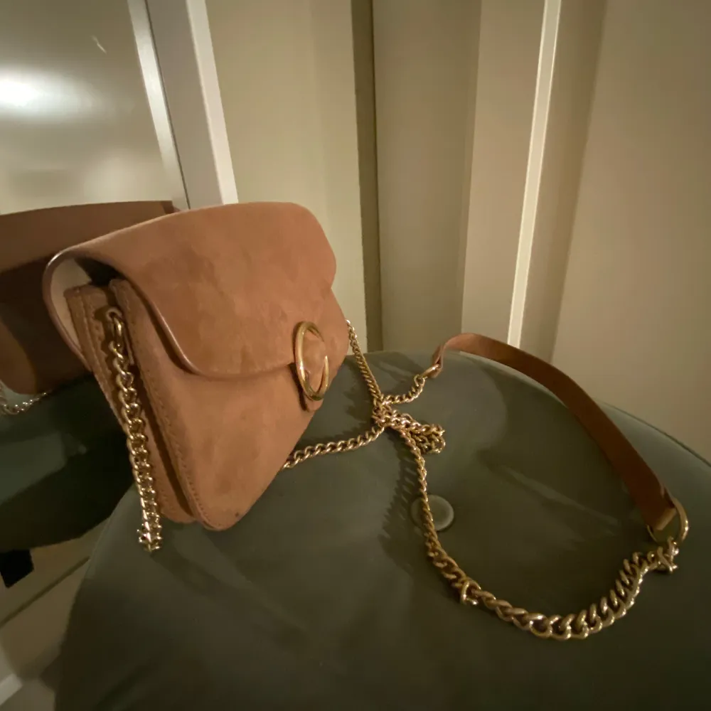 Säljer en snygg brun axelväska i mocka med en stor guldfärgad ringdetalj på framsidan. Väskan har en elegant guldkedja som axelrem och är perfekt för både vardag och fest. Den är i mycket bra skick och redo att användas direkt!. Laukut & Käsilaukut.
