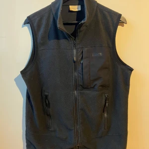 Svart fleeceväst från Wood Wood - Säljer en svart fleeceväst från Wood Wood i använt skick. Västen har en dragkedja framtill och en praktisk bröstficka med dragkedja. Den har ett minimalt hål (Vänligen se bifogad bild) därav priset. Perfekt för lager-på-lager under höst och vår. 