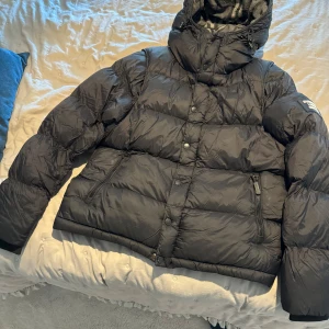 Svart dunväst med huva - Säljer en svart dunväst i mycket bra skick. Den har en quiltad design med dragkedja och knappar framtill. Västen har en avtagbar huva med ett rutigt mönster på insidan. Perfekt för höst och vinter!