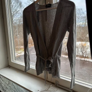 Grå kofta med knytband peak performance  - Säljer en stilren grå kofta med knytband i midjan. Perfekt för lager-på-lager under höst och vår. Den har långa ärmar och är gjord i ett mjukt material som känns skönt mot huden. Passar både till vardags och lite finare tillfällen.