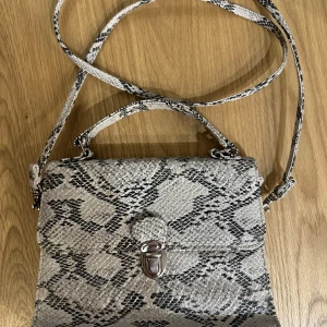 Ormmönstrad axelväska - En supercool axelväska med ormmönster i svart och vit/beige. Väskan har ett stilrent spänne framtill och en axelrem. Perfekt för att ge din outfit en edgy touch! 