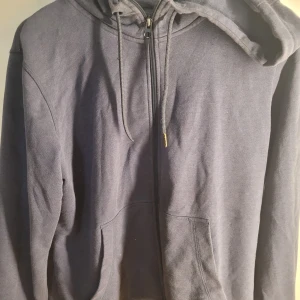 Mörkgrå hoodie från The Essentials - Säljer en mörkgrå hoodie från The Essentials i ekologisk bomull. Den har en dragkedja framtill och en skön huva med snören. Perfekt för kyliga dagar eller mysiga hemmakvällar. Passar både till vardags och avslappnade tillfällen.