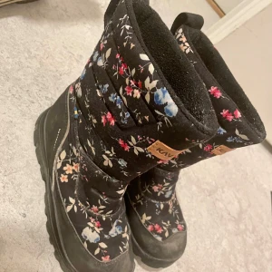 Blommiga vinterstövlar från Kavat 32 - Säljer ett par svarta vinterstövlar från Kavat med ett fint blommigt mönster i rosa, blått och vitt. Stövlarna är jätte lätta att ta på och perfekta för kalla vinterdagar, och har en robust sula för bra grepp. De är i bra skick och redo för nya äventyr i snön!