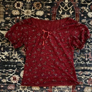 Röd blommig topp med knytband - Säljer en söt röd topp med blommigt mönster och knytband framtill som knyts till en rosett. Toppen har korta puffärmar och en bekväm passform. 🤍