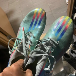 Färgglada fotbollsskor från Adidas - Säljer ett par coola fotbollsskor från Adidas i regnbågsfärger. Skorna har en snygg design med blå, rosa och gula detaljer och är perfekta för fotbollsplanen. De har snörning för bra passform och är i bra skick. Perfekta för dig som vill sticka ut på planen!