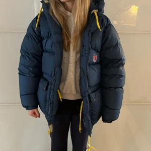 Blå dunjacka från Fjällräven - Säljer en supermysig blå dunjacka från Fjällräven i storlek XXS. Jackan är perfekt för kalla vinterdagar med sitt varma dunfoder. Den har en praktisk huva och gula detaljer som ger en snygg kontrast. Jackan är i mycket bra skick och redo för nya äventyr! 🧥❄️ Modellen är Expedition artikelnummer. 80600. ☃️Osäker om pris? Skriv så kan vi diskutera!! Äldre modell men ser ut som ny! 