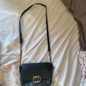 Svart axelväska med gulddetaljer - Snygg svart axelväska i skinn med gulddetaljer. Väskan har en justerbar axelrem och ett elegant spänne framtill!