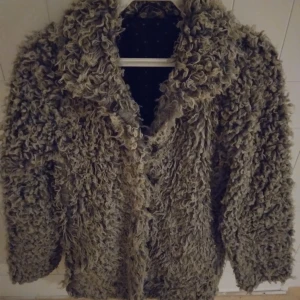 Grå fluffig jacka i fårull-still, Strlk: Small / 34-36. Fickor. - En grå fluffig jacka i fårull-stil, med mjuk och skön känsla. Den har en klassisk krage och knappar framtill, samt fickor och mörkt prickigt foder. Storlek uppskattas till Small, 34-36. Inga kända defekter.